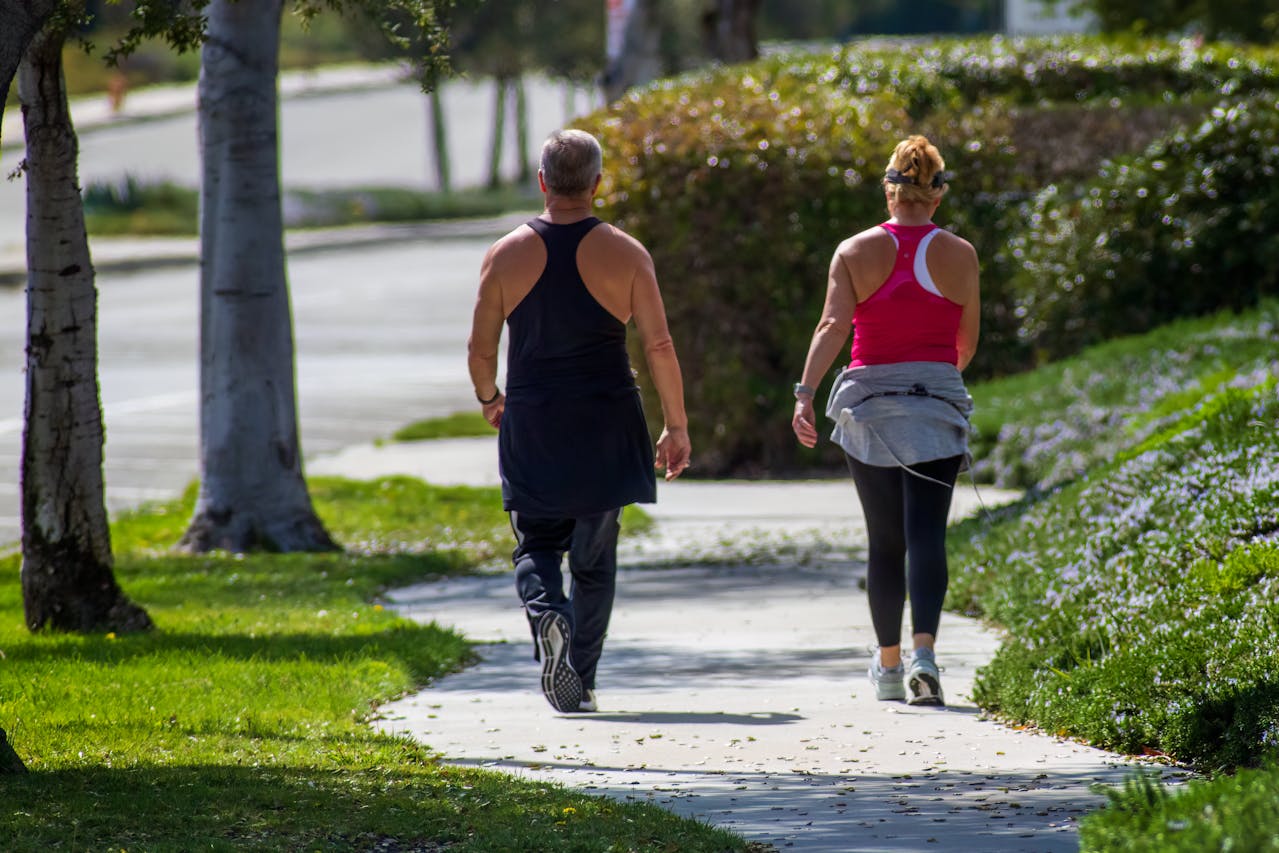 Two adults walk along a sunlit sidewalk bordered by grass and trees. The person on the left wears black athletic clothing, while the person on the right wears a pink top and has a gray sweatshirt tied around their waist.